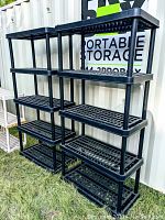 Two black plastic stackable shelving units with four shelves each, placed outdoors on grass in front of a portable storage container.