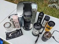 Overall shot showing Keurig K75 Platinum coffee maker with digital display, water reservoir, and drip tray. Surrounding the maker are several coffee accessories including travel mugs, a French press, and grinders.