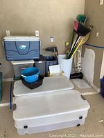 Wide shot showing blue Coleman Xtreme cooler on top of storage bins, white tall bin holding cleaning tools and umbrella, floor area with two large white storage bins stacked.