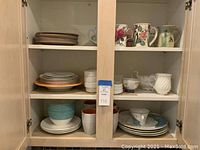 Open kitchen cabinet displaying an assortment of dishes and mugs placed on three shelves including floral mugs, white ribbed mugs, blue bowls, patterned plates and bone china teacups.