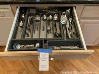 Open drawer containing stainless steel flatware organized in a grey plastic tray, visible pieces include various sizes of forks, spoons, and knives.