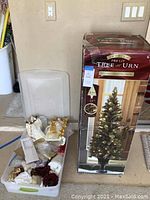 Photo of boxed pre-lit Christmas tree with urn base and storage bin of assorted holiday decor items beside it.