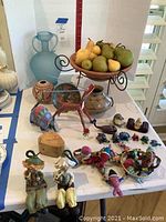 Full view of assorted decorative items including vases, wooden bowls, fruit bowl with faux fruit, fabric and wooden figurines, and two seated hanging figurines on table