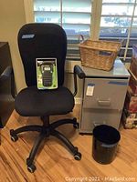 View showing black office chair with armrests, a Dymo label maker in original box on the seat, a wicker basket on top of a gray metal file cabinet with two drawers, and a small black waste bin on the floor.