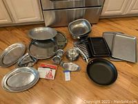 Multiple pots, pans, baking sheets, trays, and small kitchen accessories arranged on floor.