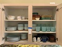 Wide view of cabinet showing multiple shelves with various stoneware dishes, mugs, and glassware arranged.