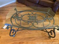 Full view of the rectangular glass top coffee table showing clear glass top and wrought metal base with scrollwork and central medallion.