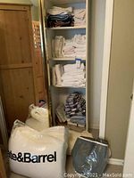 Photo of shelves with assorted folded towels, cloth napkins, and mattress topper in plastic bag labeled Crate & Barrel.