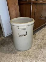 Front and side view of a large beige Western stoneware crock with metal side handles with wooden grips, placed on carpeted floor near wooden furniture.