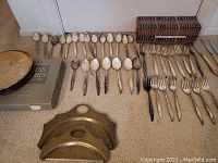 Photo of silver plated spoons, forks, knives, two boxed sets, and the floral design metal trivet laid out on carpet.