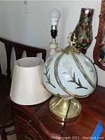 Photo showing three lamps including multicolored glass shade hurricane lamp, brass base lamp with floral globe shade, and white lamp with detached shade.
