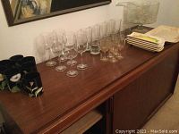 Glass punch bowl on stand with matching clear glass cups and ladle, stemware glasses, and Fitz and Floyd mugs arranged on a wooden cabinet.
