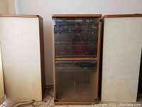 Full view of the stereo system cabinet flanked by two floor-standing speakers with beige fabric covers and light wood trim.