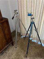 Photo showing two tripods standing on a floor, one silver, one blue, in front of curtains and next to wooden furniture.