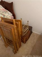 Four wooden TV trays folded and arranged in a stand next to a bed, with a wooden doll cradle, a picnic basket, and a wooden box nearby.