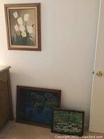 Framed white tulip painting on wall above two smaller nature paintings leaning against furniture