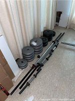 Photo showing multiple grey weight plates in different sizes stacked against the wall alongside three bars (one full-length chrome barbell bar and two shorter dumbbell bars).