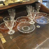 Six clear glass stemware glasses and five clear glass dessert plates arranged on a wooden surface, showing the full set including the beaded details.