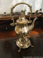 Full view of antique brass tea pot showing ornate handle, domed lid, curved spout and attached to footed pouring stand with curved legs.