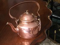 Copper kettle placed on a polished wood table, showing full side view including curved spout and handle.