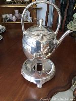 Front and side view of the silver-plated tea pot on its warming stand sitting on a wooden table, showing the curved spout, hinged lid, and handle design.