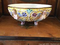 Front left angle showing three-dimensional fairy faces on bowl sides and floral painted decoration on bright yellow background, porcelain material, gold rim detail.