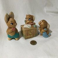 Three Pendelfin bunny figurines arranged around a coin for scale: standing bunny, bunny at desk, and crying bunny holding a polka-dot blanket.