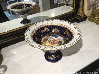 Angled view of the cobalt blue vase sitting on a marble surface showing the broad scalloped rim with gilt decorations and the base reflecting in a mirror.