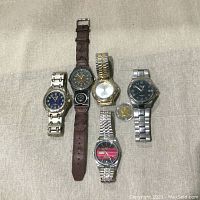 Group photo showing five men's wristwatches with varied bands: metal and leather. Watches arranged on a beige fabric surface. Includes a Canadian toonie coin for scale.