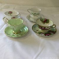 Four distinct floral porcelain cups and saucers arranged in two rows on white fabric background.