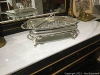 Full view of silver plate entree dish with lid on warmer tray placed on marble surface with mirror background.