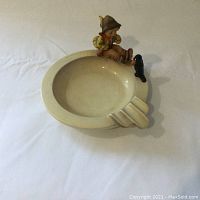 Full view of cream-colored porcelain ashtray with boy and blackbird figurine on rim.