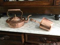 Kettle and water pourer displayed side by side on marble surface in front of dark wood furniture