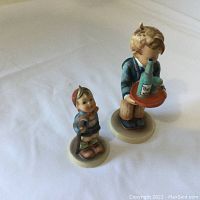 Two Hummel figurines displayed on white background, the taller boy holding a tray on right, smaller boy with a stick on left.