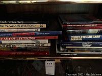 Photo of a collection of automotive themed hardcover books on a wooden shelf, titles visible and stacked horizontally and vertically.