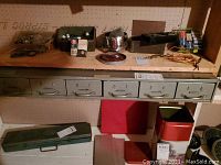 Straight on view of a long metal cabinet with seven drawers filled with various small parts and tools laid out on a shelf. Surrounding shelves contain tool boxes, containers, and other hardware items.