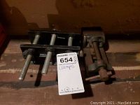 Three heavy duty metal clamps shown on a wooden surface. Two have multiple bars and threaded rods; one is a single screw clamp.