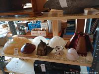 Various vintage lamp shades on shelves including glass, ceramic, metal, and fabric types along with a small metal lamp base, showing condition and variety.