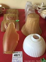 Photo showing six of the vintage glass lampshades on a red surface, including amber elongated, ribbed white, bell-shaped beige, and three cream square ones with embossed patterns.