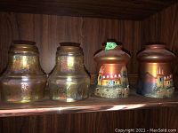 Four glass light shades on a wooden shelf, two are iridescent yellowish with gold floral decoration and two are darker with hand painted building scenes. One chipped top edge shade is visible