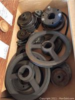 Box with a variety of metal pulleys of different sizes and shapes, some with grooves and mounting holes, showing surface rust and wear.