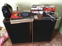 Pair of large speakers flanked by stacks of vinyl records, cassette tapes, VHS tapes, and a Linksys router/cable modem on top