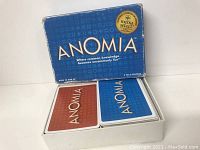 Blue rectangular Anomia game box with gold and white text. Box top is viewed partially open, showing two decks of cards inside.