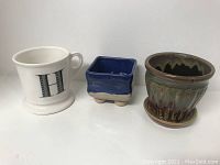 Photo showing one white ceramic mug with letter 'H', one small blue square ceramic pot, and one round brown-green glazed ceramic pot with saucer.