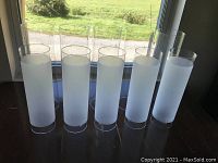 Five cylindrical glass lights with frosted lower half, clear upper half, standing upright near window.