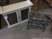 White wooden medicine cabinet with two etched glass doors and small metal knobs next to a black metal wire magazine rack with spiral design.