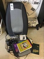 Photo of black and gray Homedics Shiatsu massaging cushion standing against a chair with health books and a hair dryer on the floor.