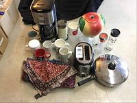 Overall view of lot items on countertop including coffee maker, toaster, frying pan, cookie jar, mugs, small bowls, salt and pepper shakers, fabric tablecloth folded in front.