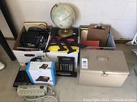 Overview photo showing a globe, file bins with clipboards and folders, metal lockbox, calculator, surge protector, and other office supplies and electronics.