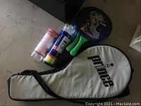 Photo showing white Prince tennis racket carrying case, two cans of tennis balls (one Wilson), and two Corfu paddles with green handles.